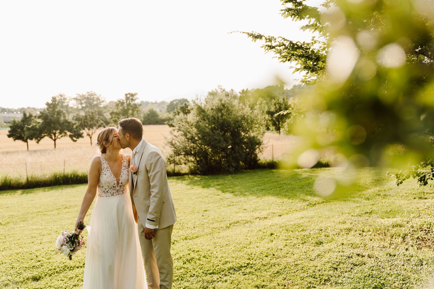 Newlyweds sharing a tender kiss framed by lush green foliage in a sunlit countryside setting.