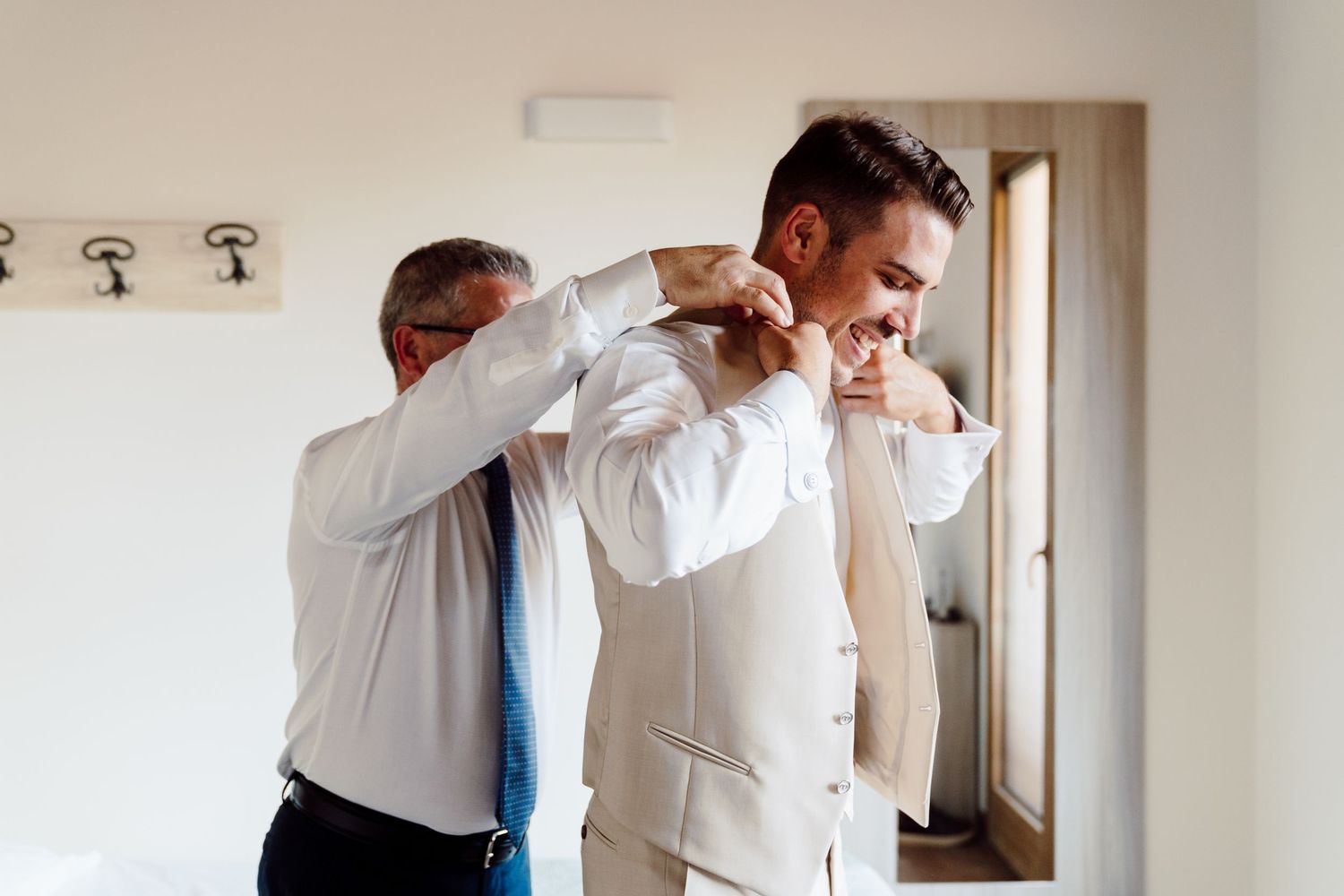 Groom getting help adjusting his white suit jacket from an older man while smiling in a bright room.