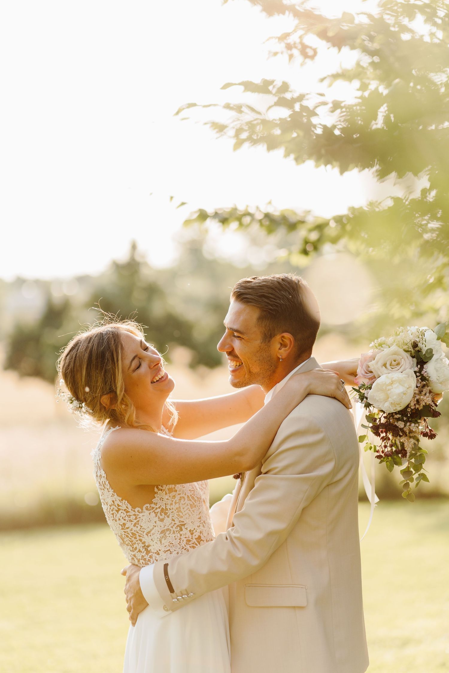 Happy bride and groom embracing outdoors in golden sunlight, bride holding a white floral bouquet.
