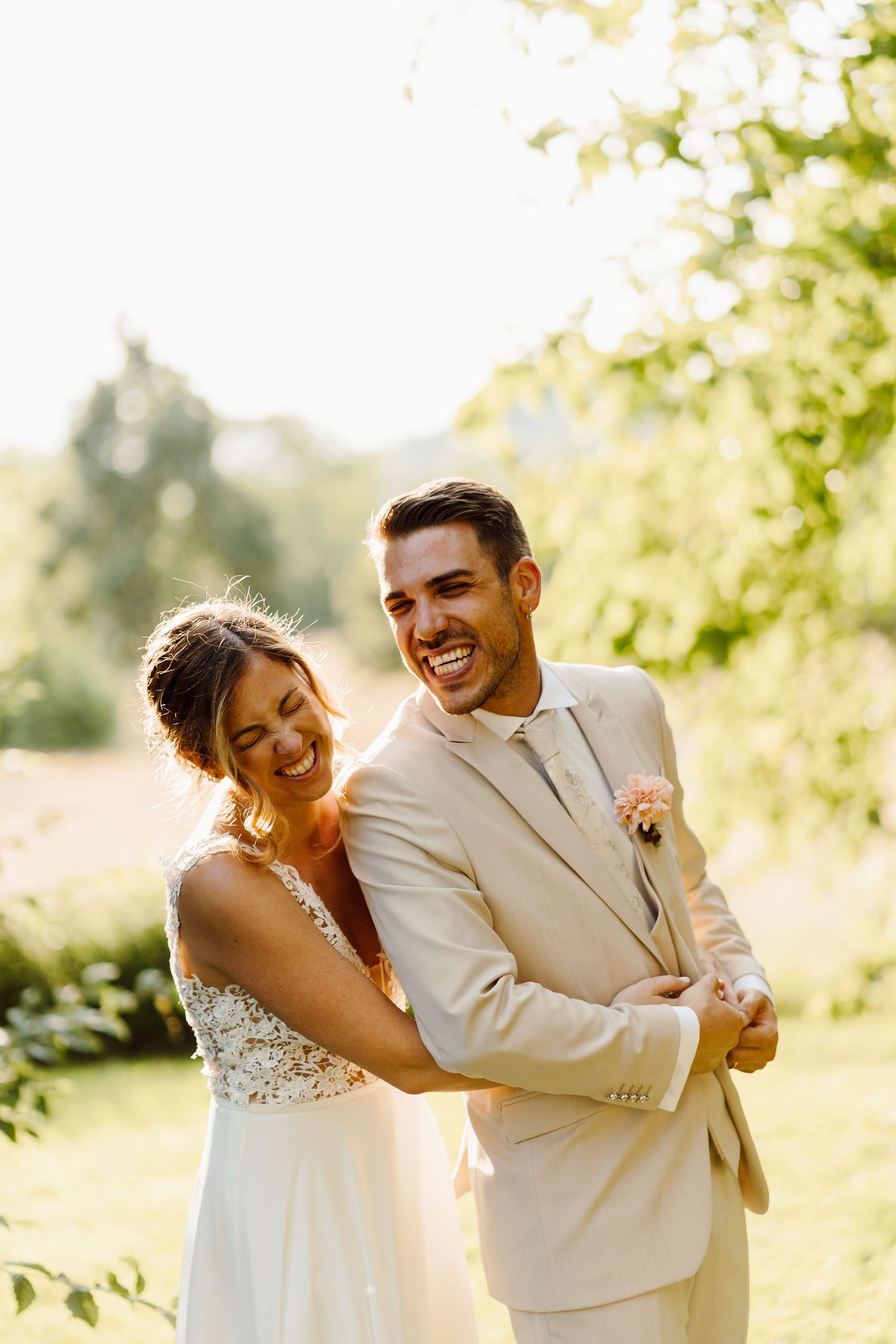 Laughing bride hugging groom from behind in a sunny garden, groom wearing a beige suit.