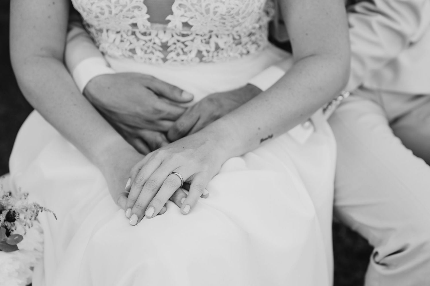 Black and white close-up of bride and groom holding hands, wedding ring visible on bride's finger.
