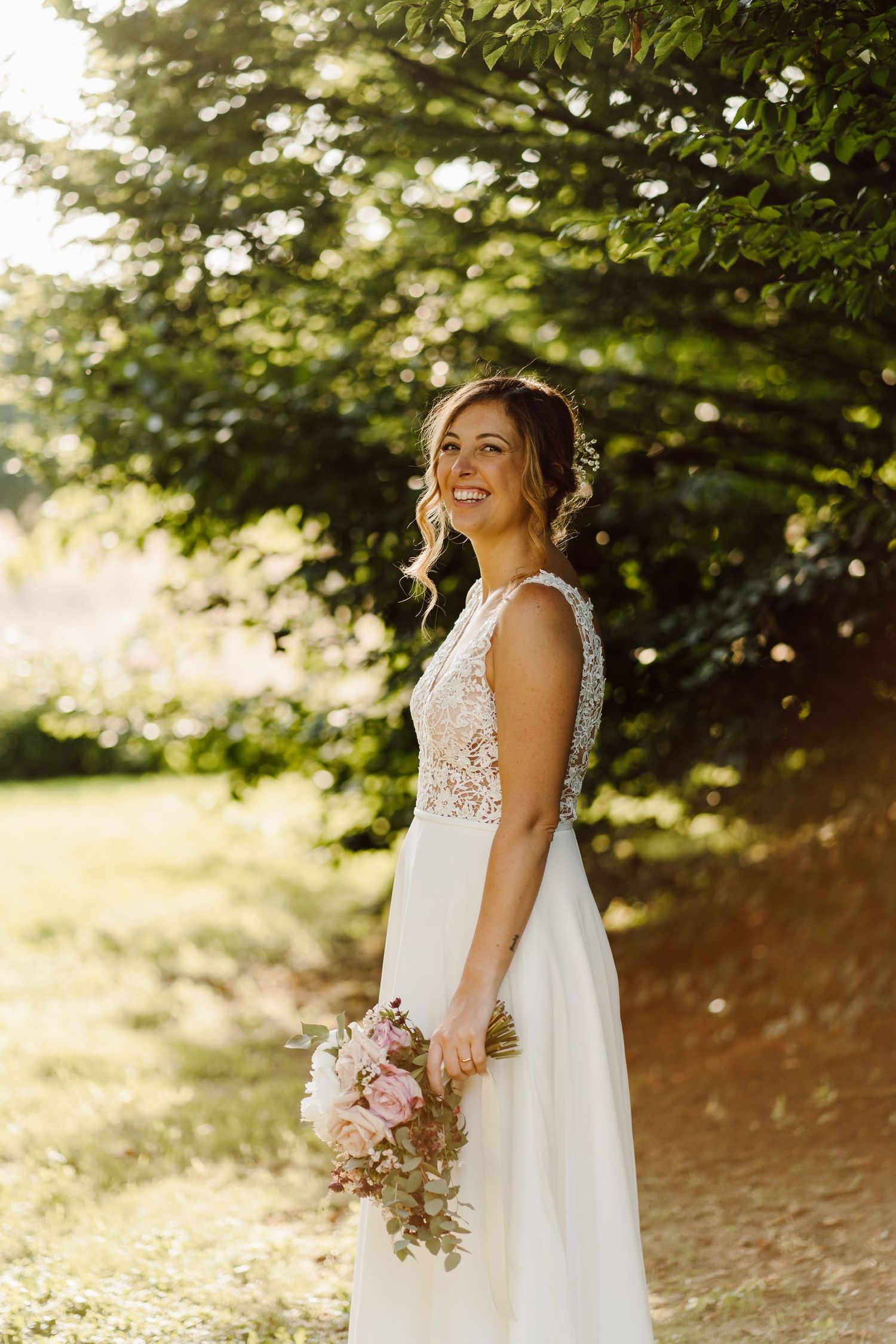 Radiant bride in a lace wedding gown smiling while holding a pink bouquet in a sunlit garden.