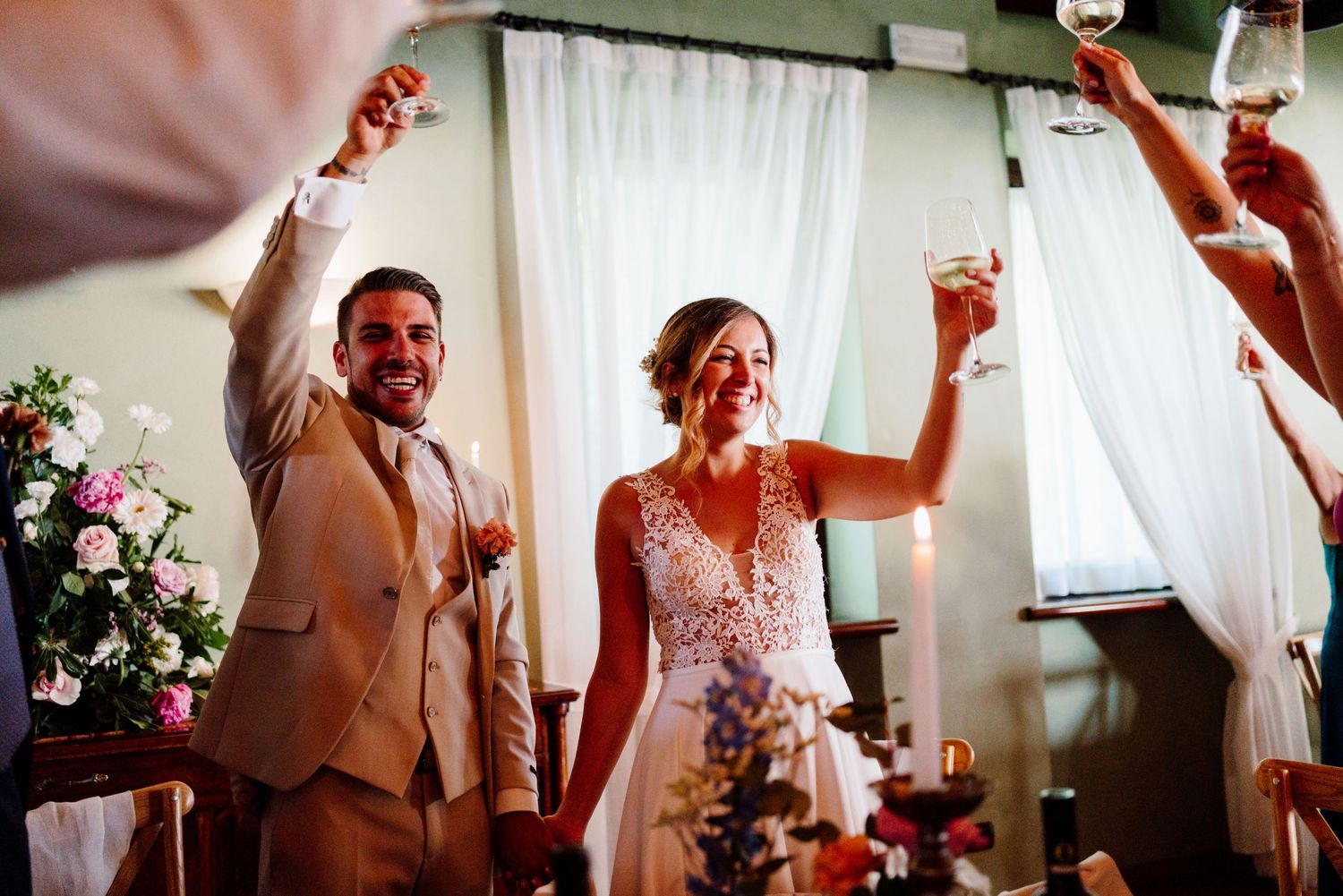 Bride and groom raising glasses in a joyful wedding toast surrounded by guests and floral decorations.