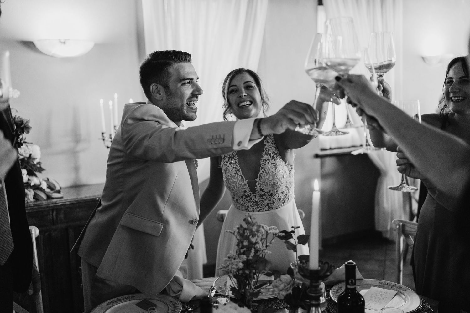 Black and white photo of bride and groom laughing and clinking glasses during wedding reception toast.
