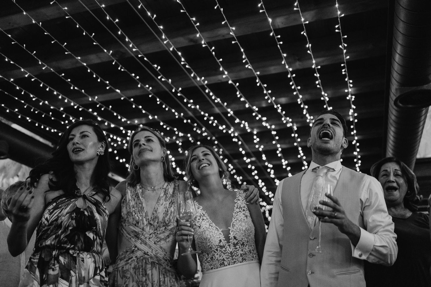 Black and white photo of wedding guests singing joyfully under a canopy of twinkling fairy lights.
