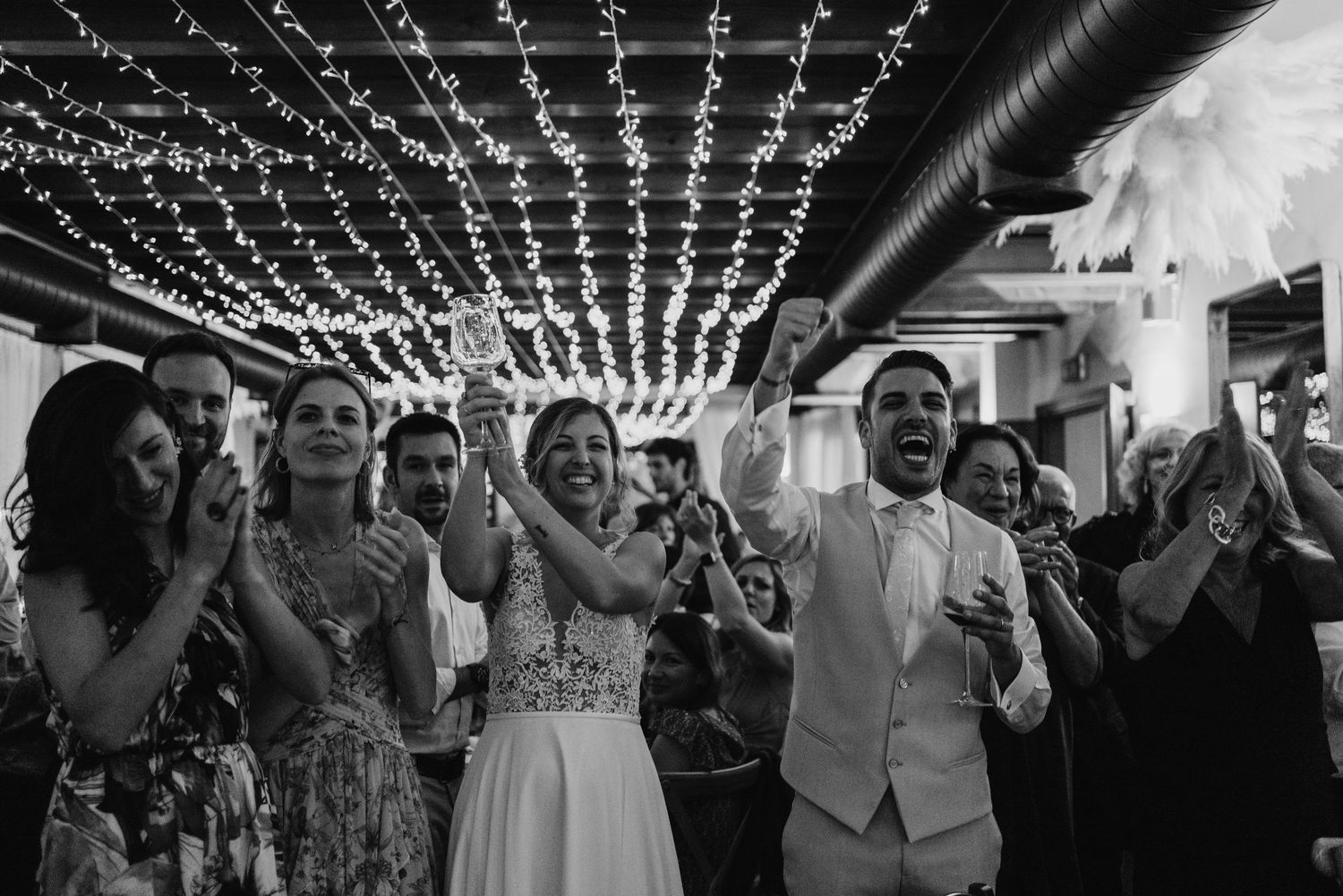 Bride and groom celebrating joyfully with guests under sparkling fairy lights at wedding reception.