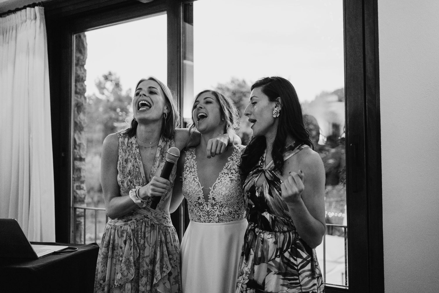 Black and white photo of bride and two bridesmaids laughing and singing together with a microphone.