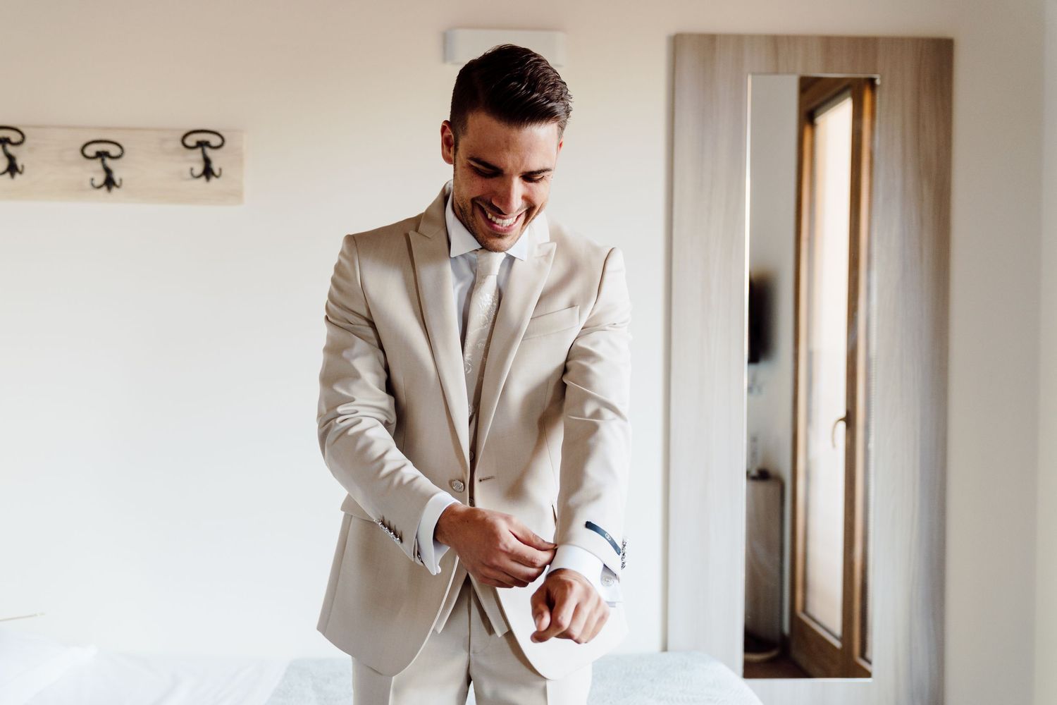 Handsome groom in a beige suit smiling while adjusting his cufflinks in a bright hotel room.
