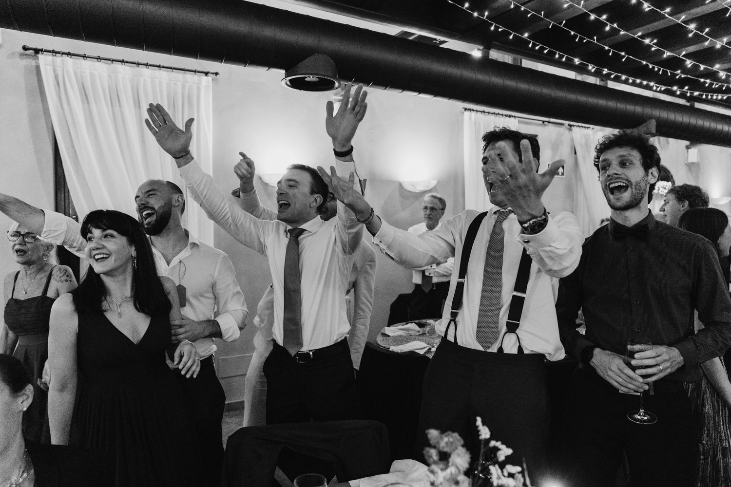 Black and white photo of excited wedding guests cheering and dancing at a lively reception party.
