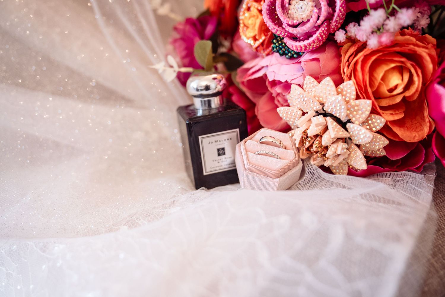 Wedding rings rest on a white surface surrounded by pink and red roses and perfume bottles.