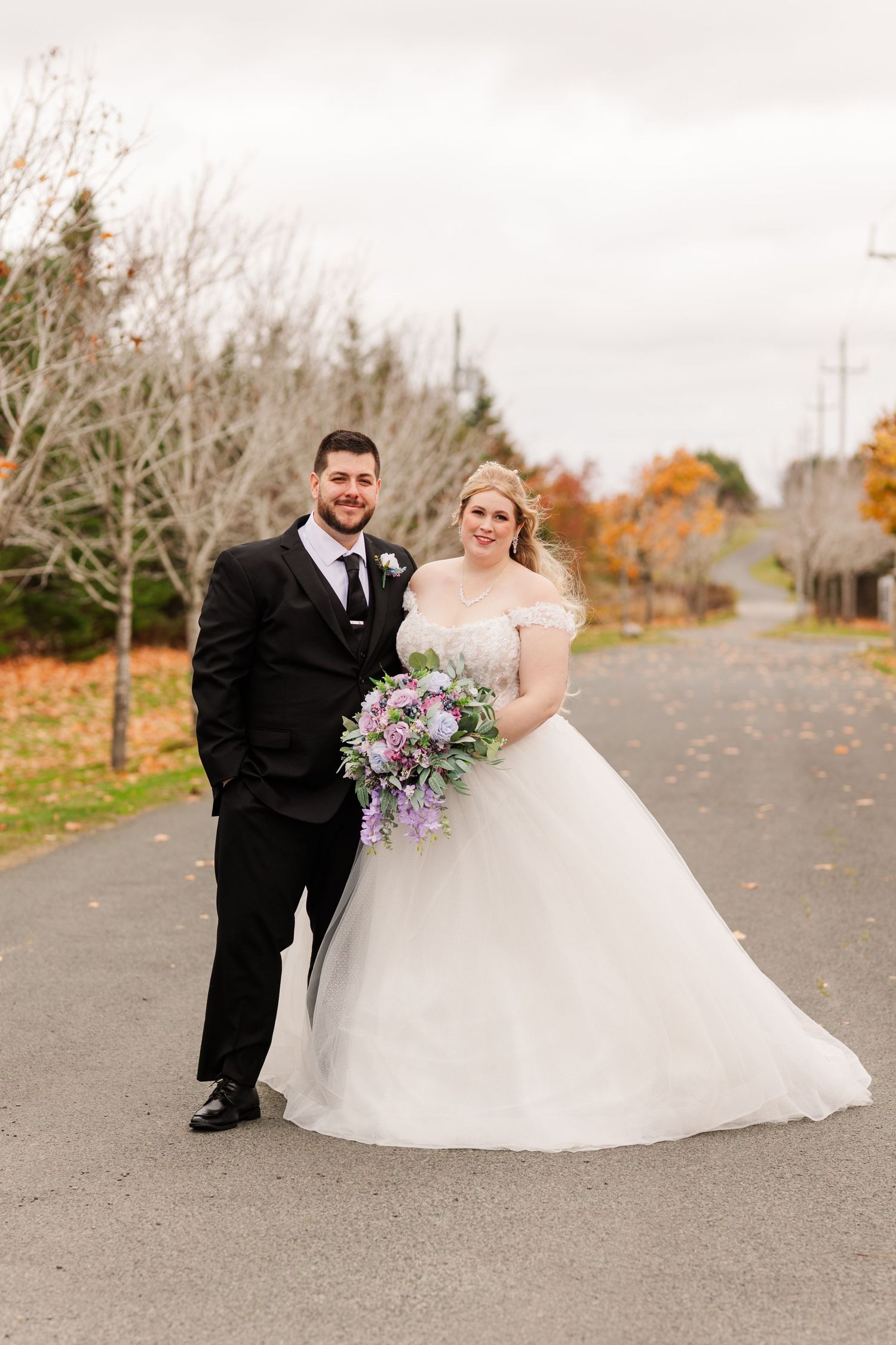 A couple in wedding attire stands together on a paved road during autumn with trees in the background.