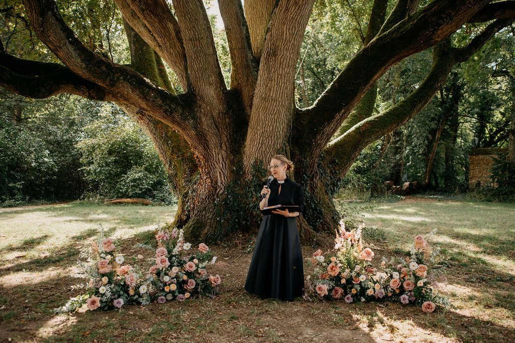 Wedding officiant in black dress stands before a grand tree with pink floral ground arrangements.