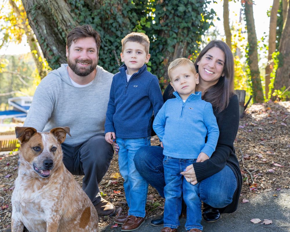 A happy family poses outdoors in a wooded area with their brown and white dog on a sunny day during family photography session in Stow, MA.