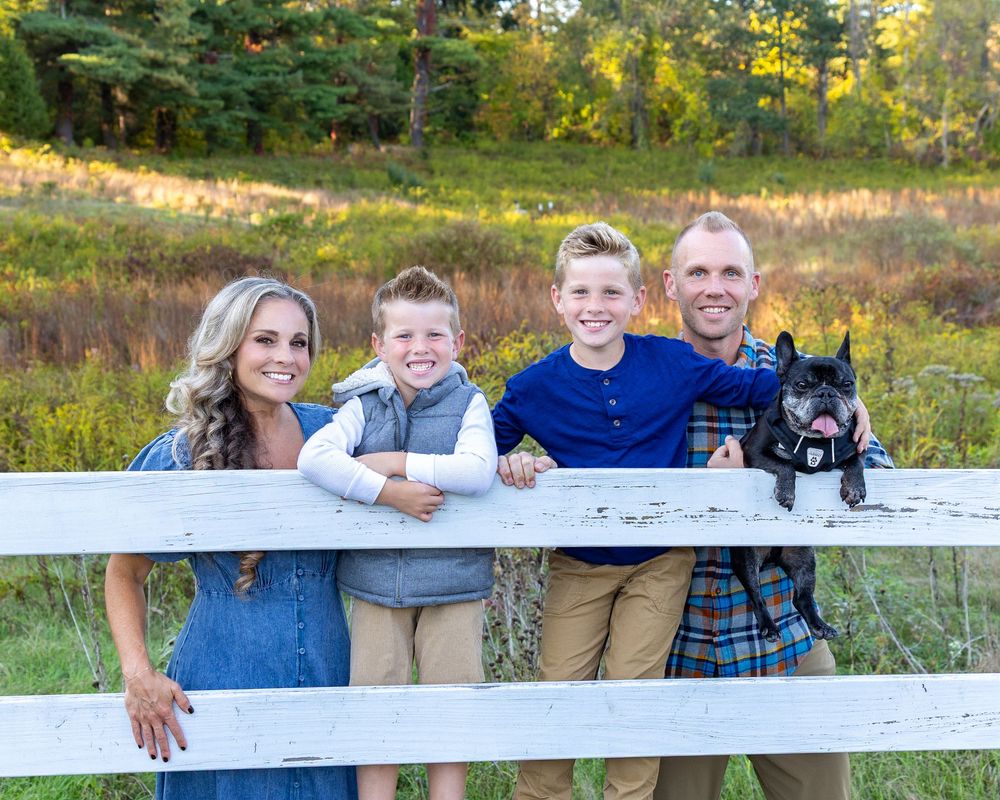 A family stands together at a white fence in a scenic autumn field with their black French Bulldog for family photo session in Sudbury, MA
