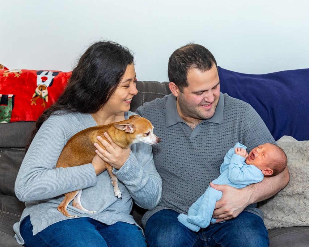 A family and pet dog gaze lovingly at a yawning newborn in a blue onesie during their newborn portrait session in Boston, MA.
