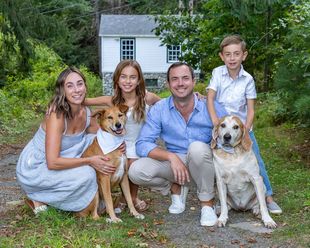 A family in coordinated blue outfits poses outdoors with their two dogs in front of a white garden shed on a summer day.