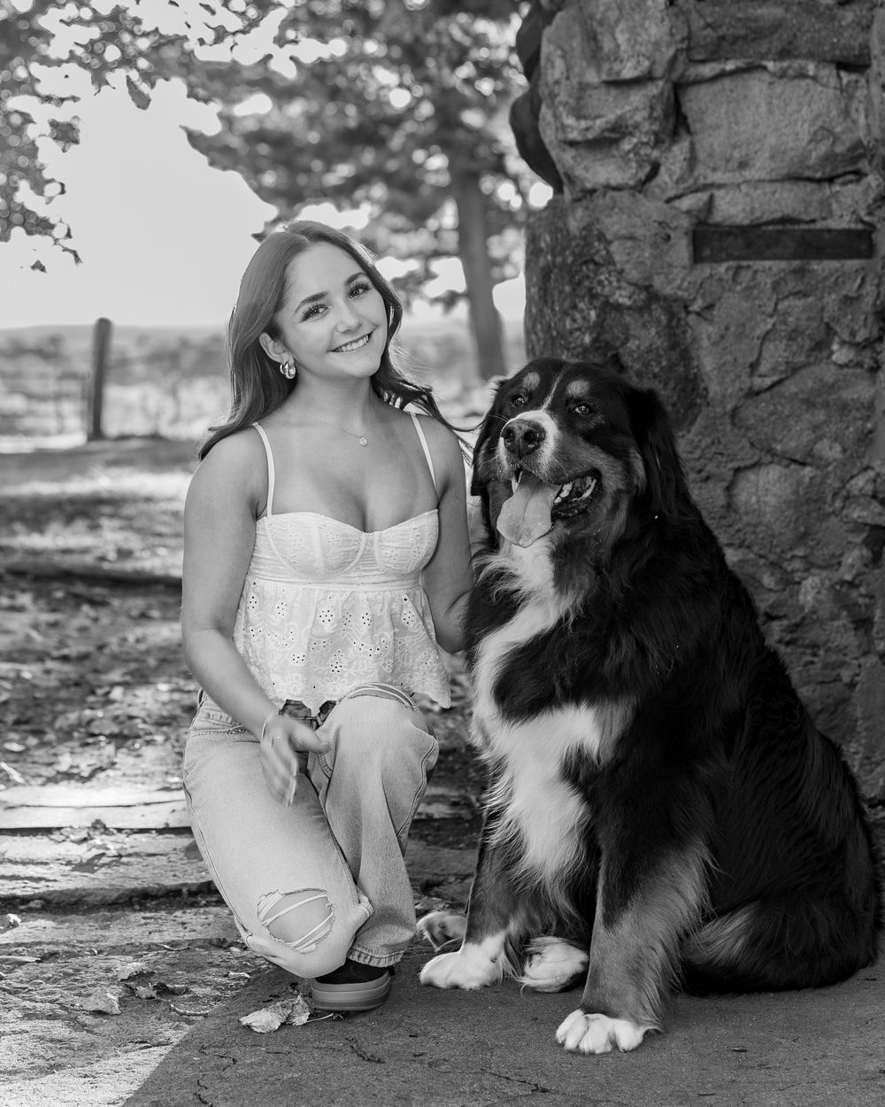 Black & white photo shows high school senior in a white dress sitting with a Bernese Mountain Dog against a stone wall at Bancroft Castle.