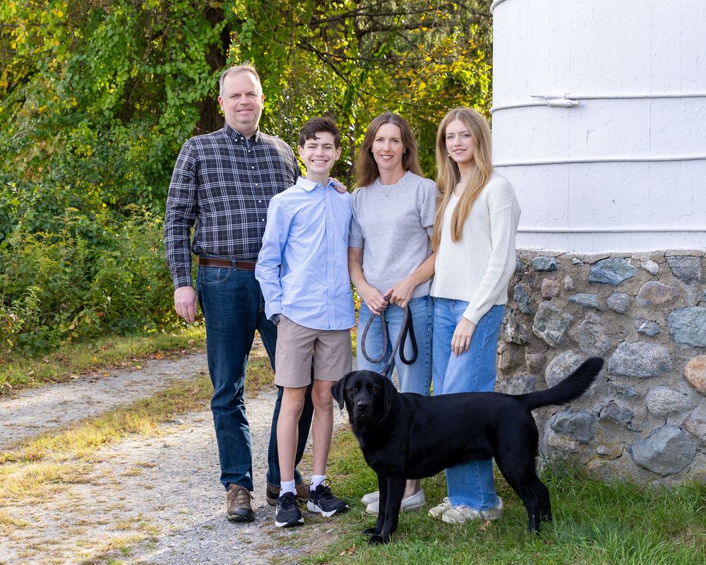 A family poses casually outdoors against a white barn & stone foundation on a sunny autumn day during a family photo session in Sudbury.