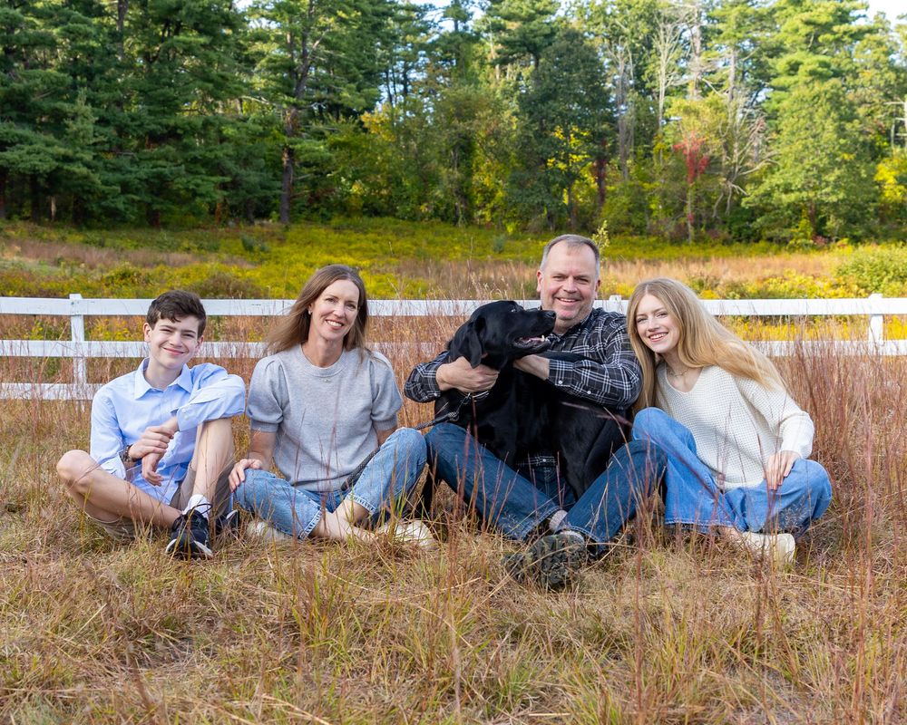 A family and their dog sit in tall grass during a casual outdoor family photography session in autumn at Wolbach Farm Sudbury, MA.
