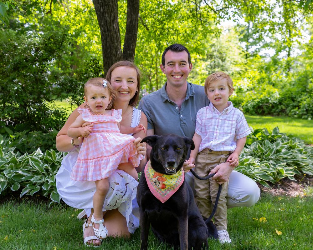 Family and their black dog pose together on grass in a lush green garden setting during a Spring family photo session in Sudbury, MA.
