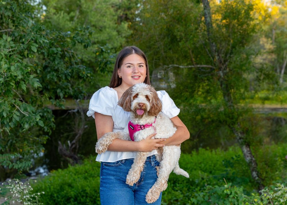 High school senior holds a brown and white dog while standing in a lush green garden setting along the Concord River for yearbook portraits.