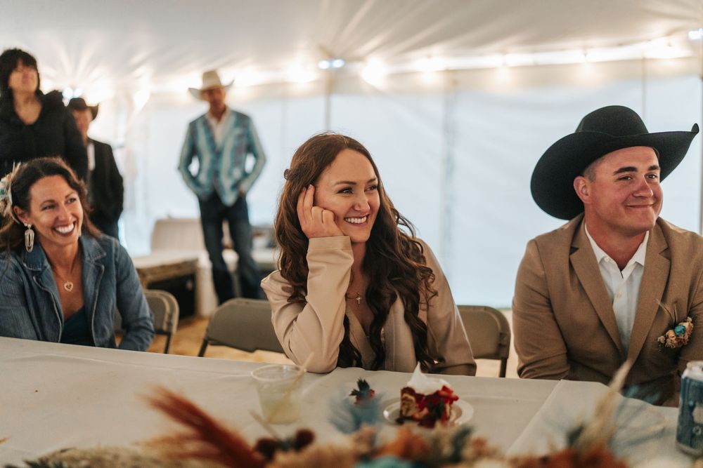 Wedding guests share laughter and conversation while seated at long tables under a white tent during a reception.