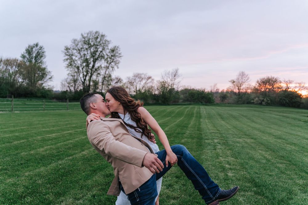 A couple embraces playfully in a green field at sunset with trees in the background.