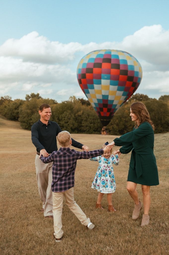 A family plays together in a field during golden hour with a hot air balloon in the background.
