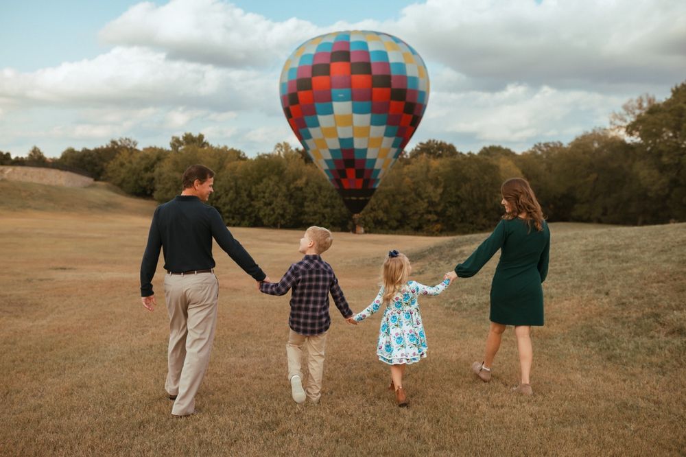 People holding hands walking through a grassy field with hot air balloons visible in the distance.