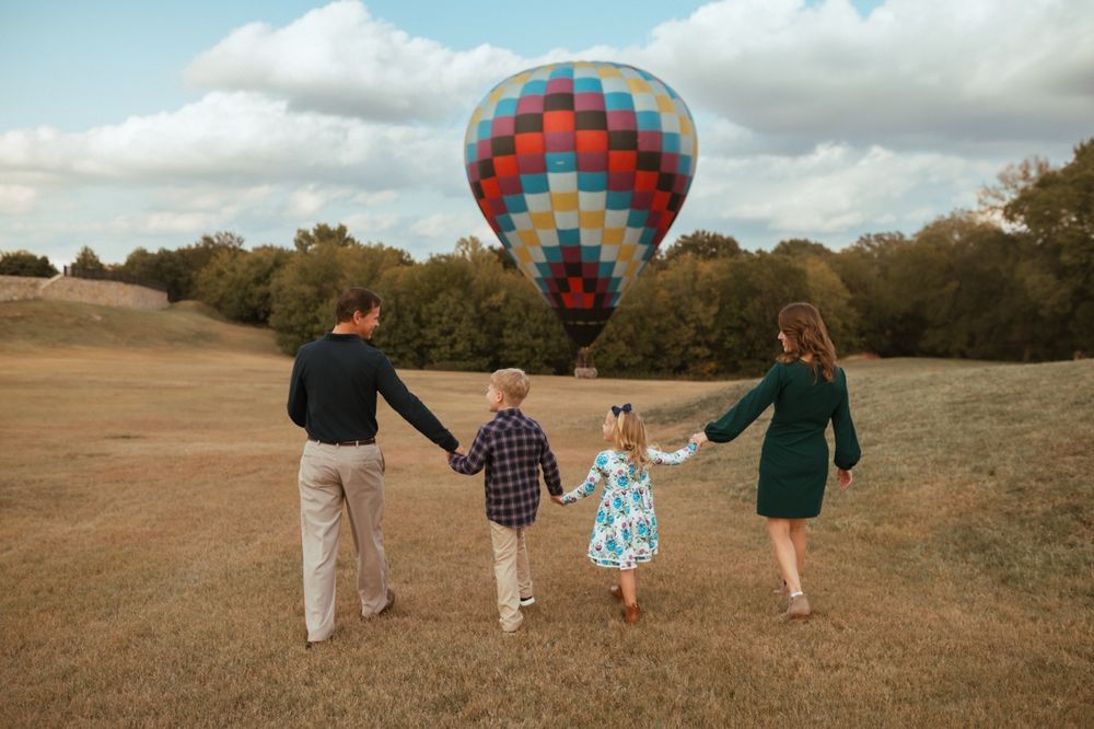 Family members hold hands while walking through a field with hot air balloons in the background.