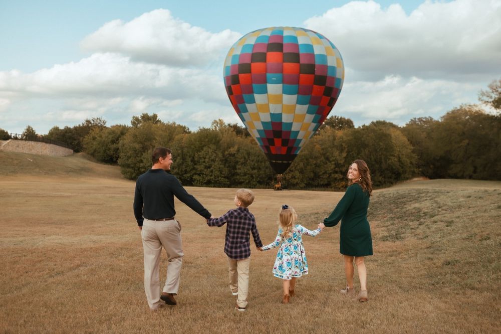 A family walks together across a grassy hillside with a vibrant hot air balloon floating in the background.