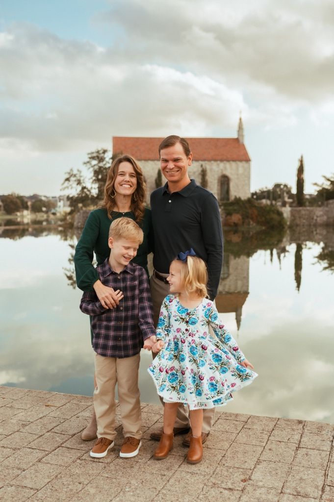 Family stands together on stone pavement by a lake during cloudy day wearing casual attire.