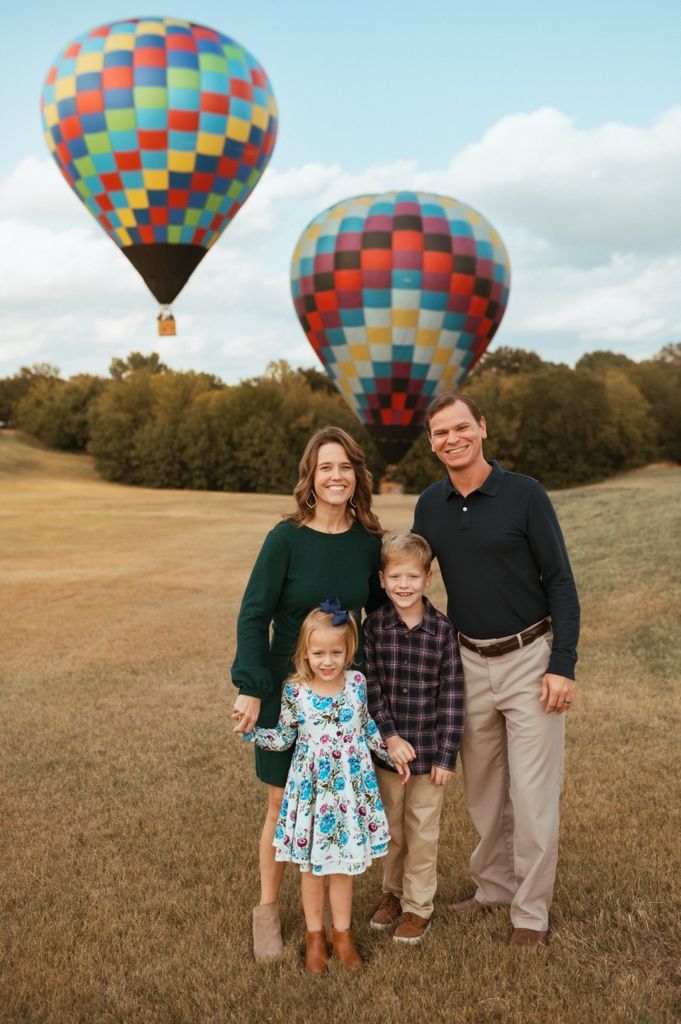 A family poses in a field with a colorful hot air balloon floating in the background on a cloudy day.