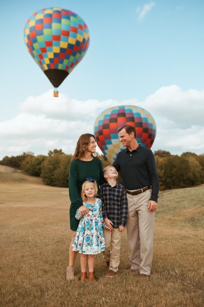 A colorful hot air balloon floats in a blue sky above a grassy field where a family gathers for photos.