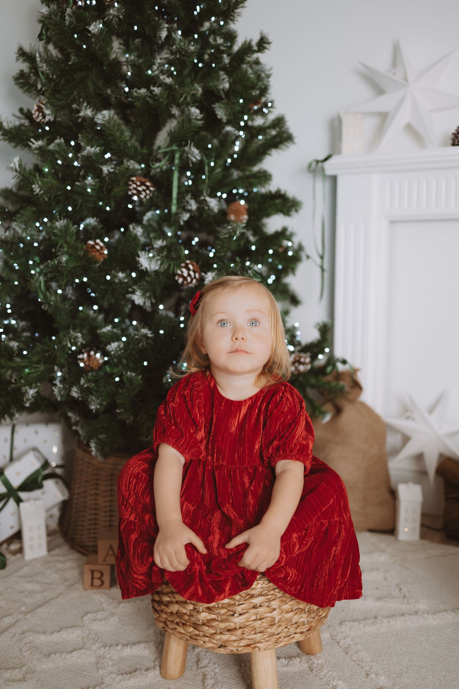 A small child in a red velvet dress sits in a wooden basket near a decorated Christmas tree.