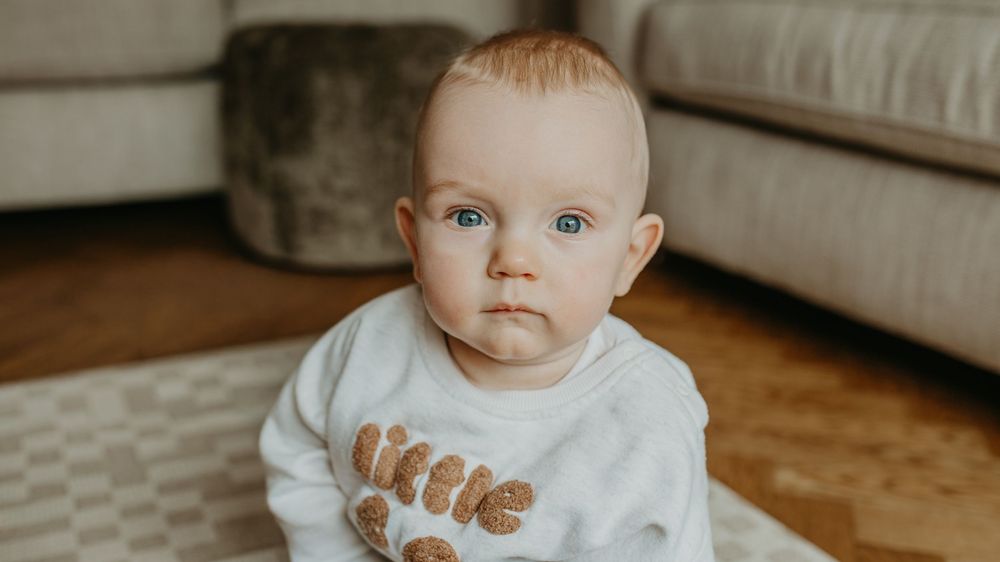 Baby wearing a white onesie sits on hardwood floor with patterned rug visible in background.