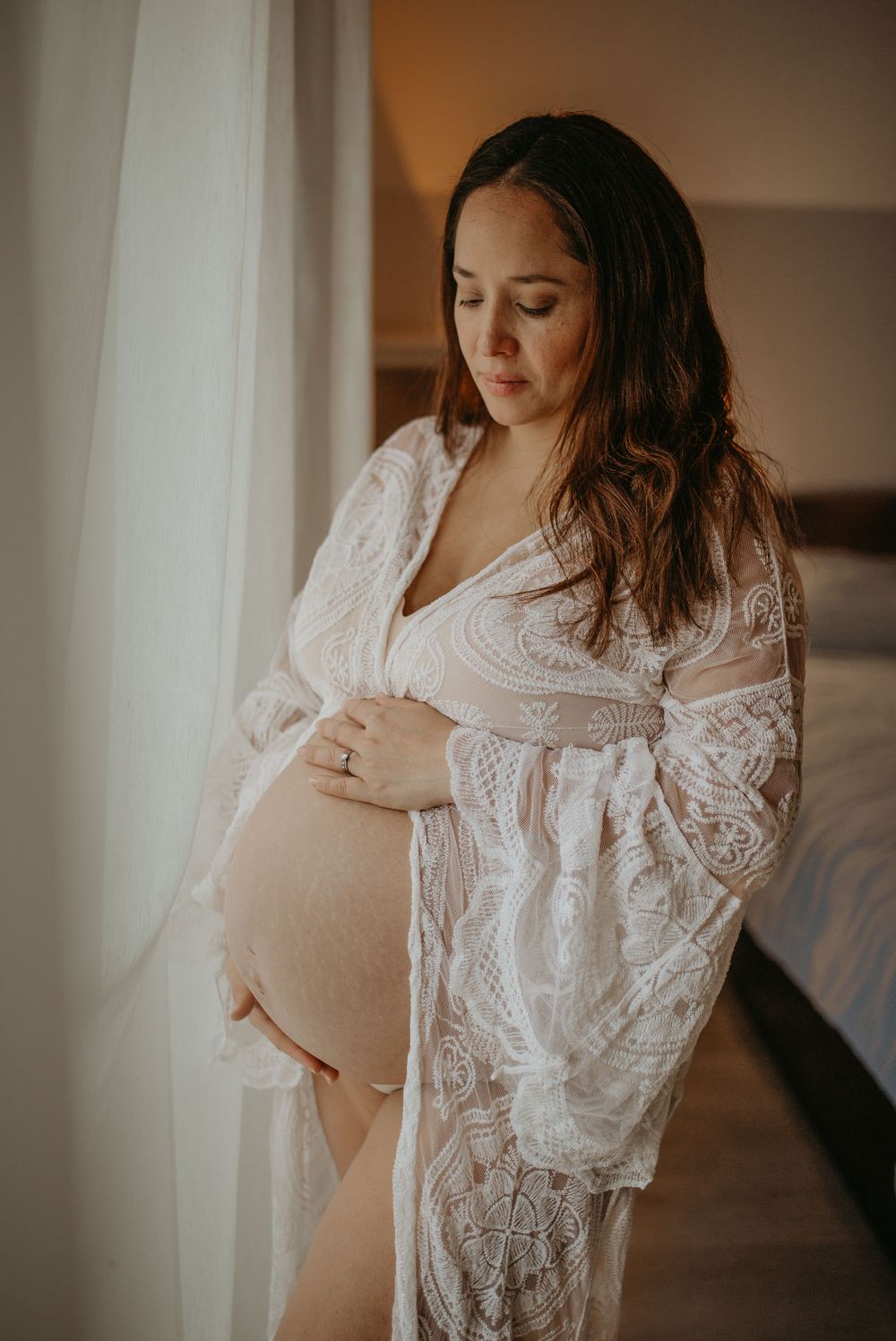 Artistic maternity boudoir photo series showing a pregnant woman in white lace robe against warm indoor lighting.