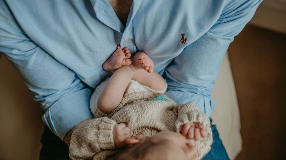 A baby wearing a light blue shirt lies on a soft cream-colored surface.