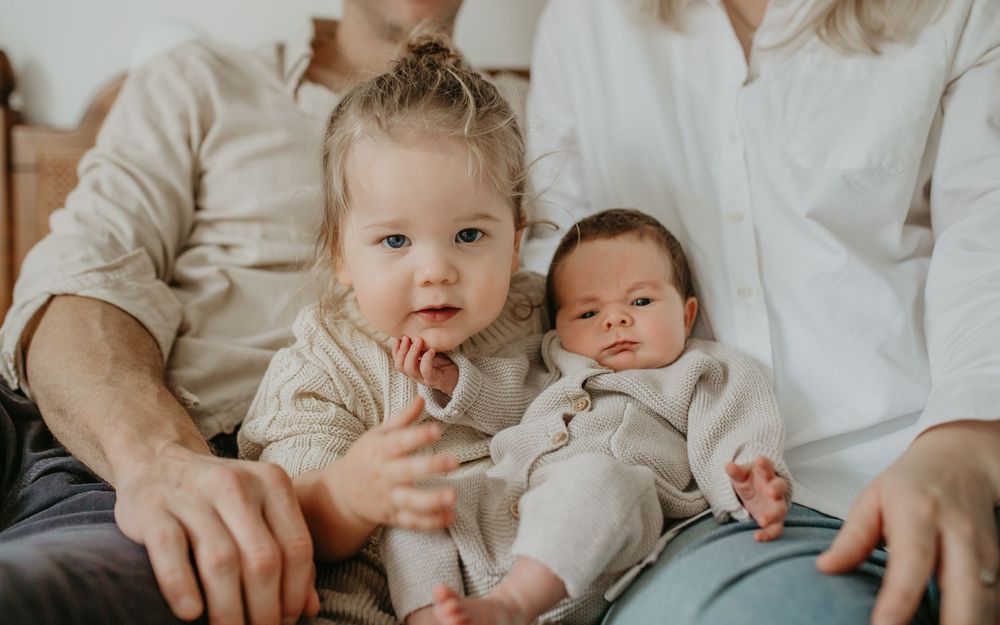 Two young children in light colored clothing sit together with their parents on a couch in a warm-toned setting.