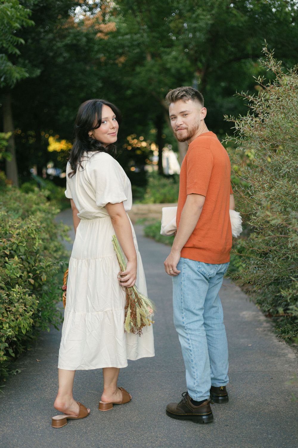 Couple in casual attire looking back at camera while walking on garden path.
