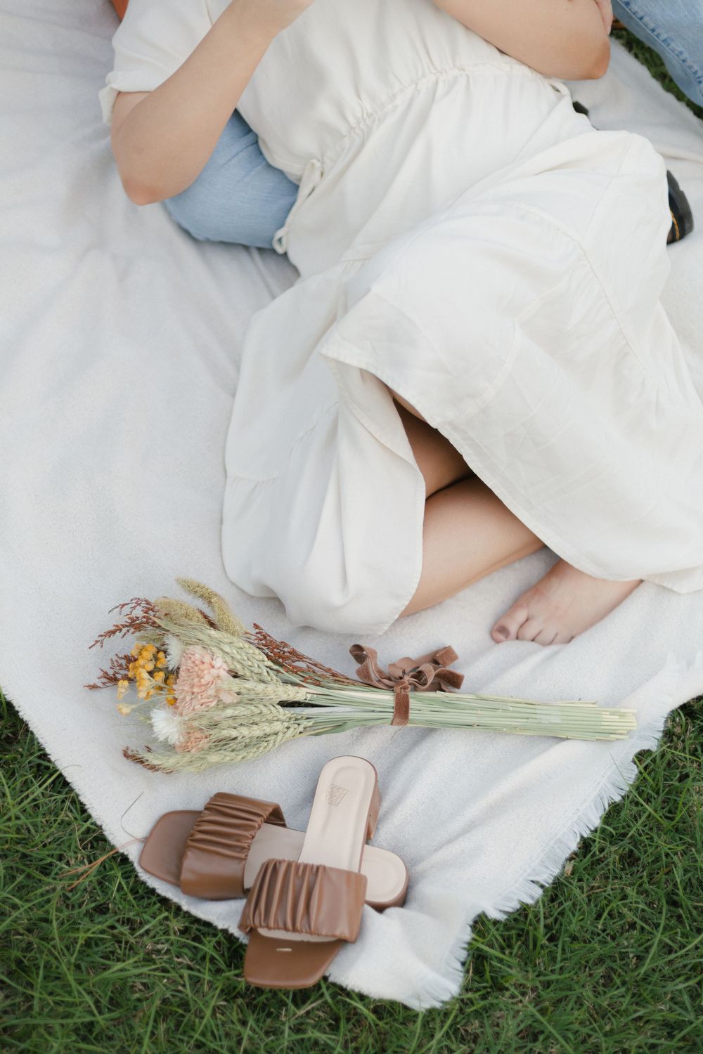 Picnic setup with dried flowers and brown sandals on white blanket on grass.