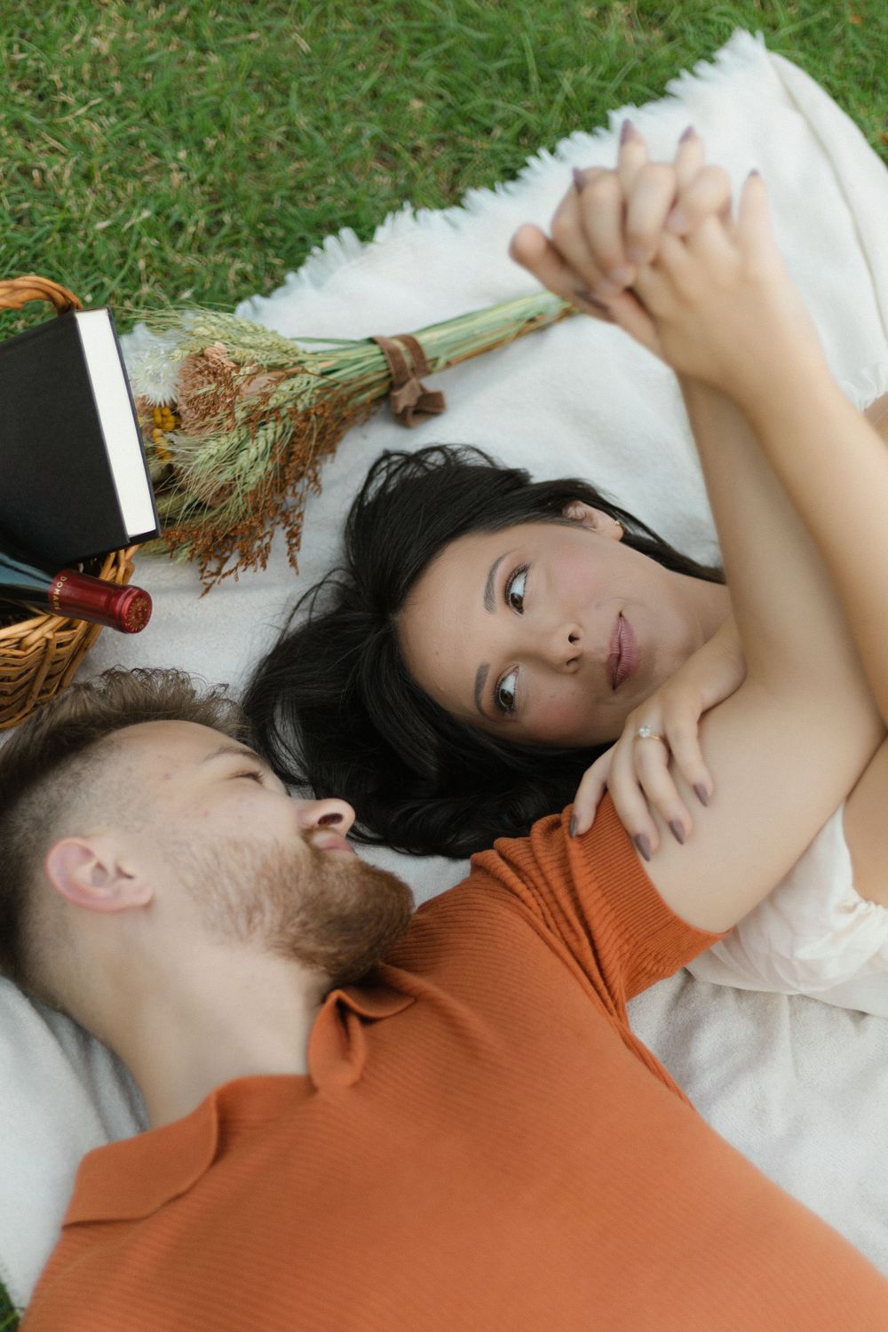 Couple lying on white blanket in grass with dried flowers, wearing orange and white outfits.