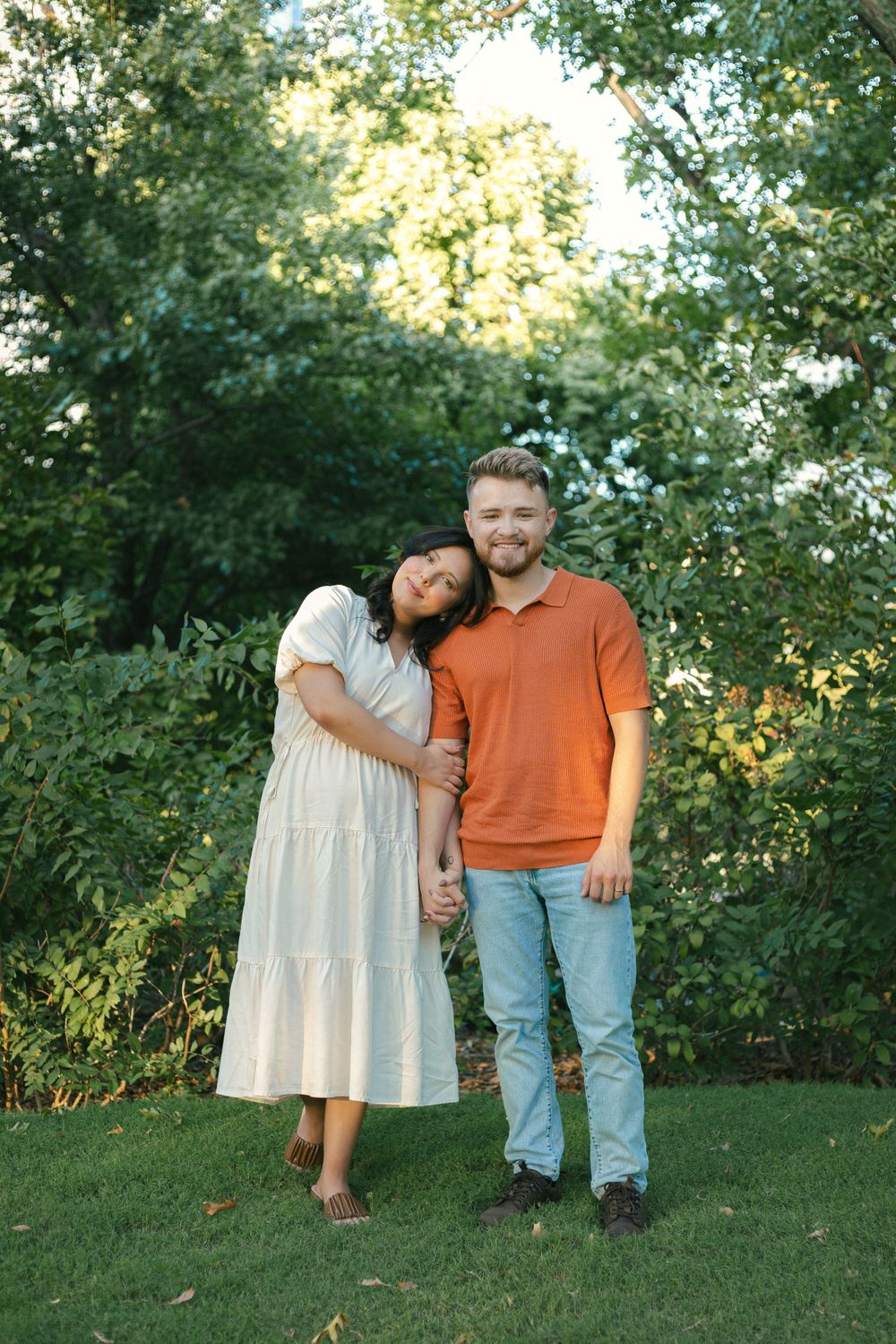 Couple in orange polo and white dress posing together on grass with trees in background.
