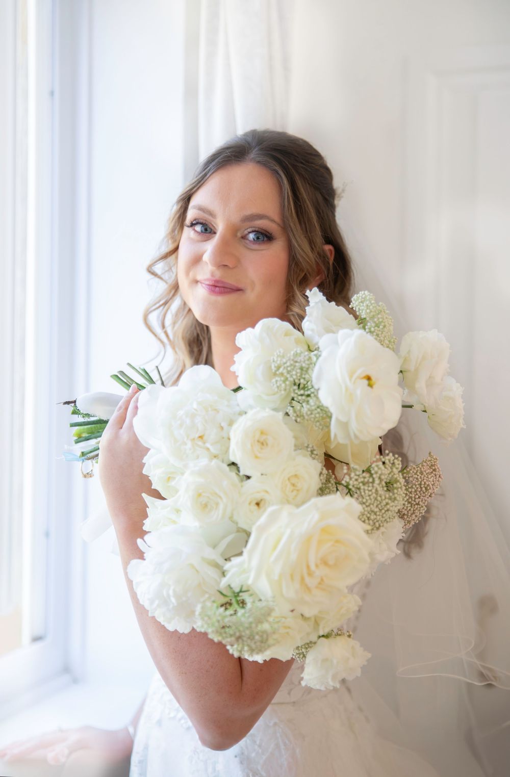 Elegant white peony and rose bridal bouquet held against a bright window backdrop.