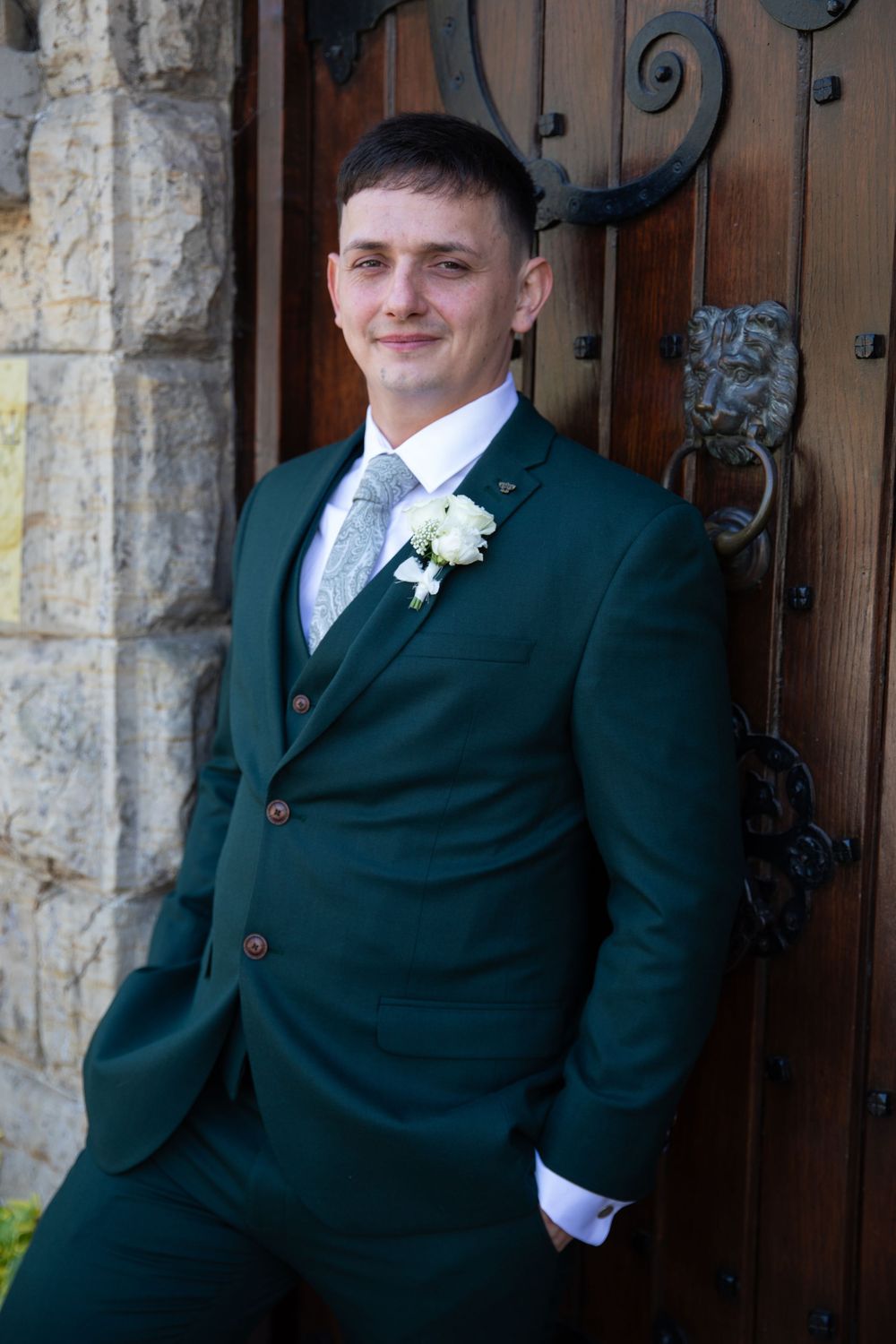 Groomsmen in dark suits posing against stone wall and wooden door.