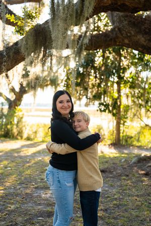 Two people share a playful hug under a sprawling oak tree in a sunlit park.