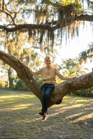 Person in beige sweater sitting on a large tree branch in a sunny park.