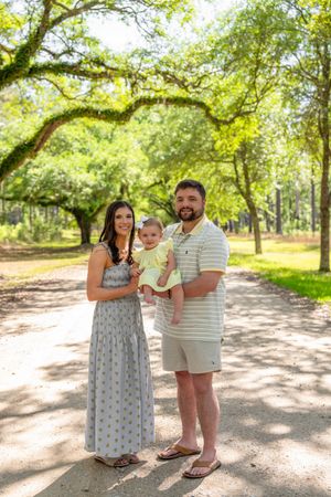 Young family with baby posing on a tree-lined path in a lush green park on a sunny day.