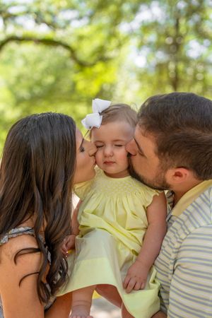 Parents kissing their baby girl on the cheeks outdoors, baby wearing yellow dress with white bow.