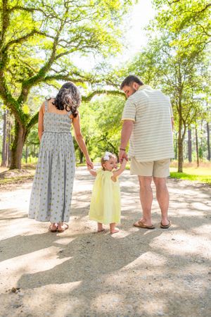 Family of three holding hands on a sunny tree-lined path, parents in casual attire with toddler in yellow dress.